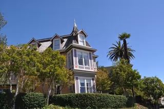 A Victorian-style senior living building with manicured hedges and palm trees under a blue sky.