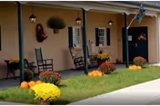 A covered porch with rocking chairs, potted mums, and pumpkins in front of a beige facility.