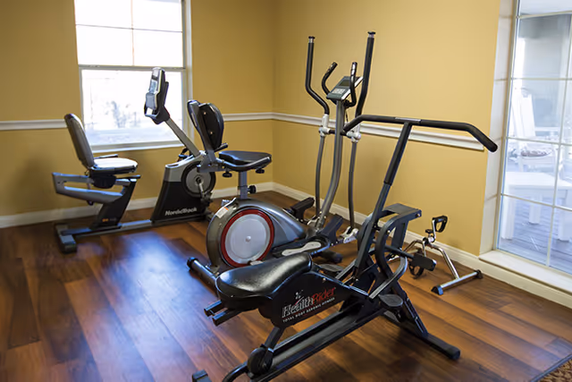 A small fitness room with wooden flooring and yellow walls, containing exercise equipment including a recumbent bike, an elliptical machine, and a rowing machine. Large windows allow natural light to enter the room.