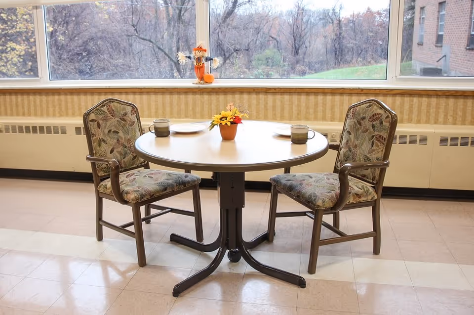 Round table with two upholstered chairs set with mugs, plates, and a small centerpiece by a large window overlooking trees.