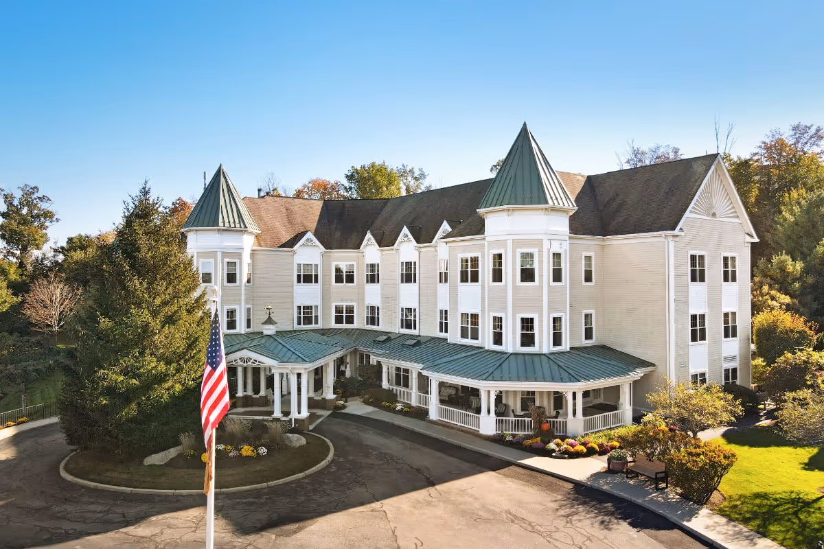 Front exterior of a multi-story senior living building with turret towers, a covered green-roof entrance, and an American flag in the driveway.