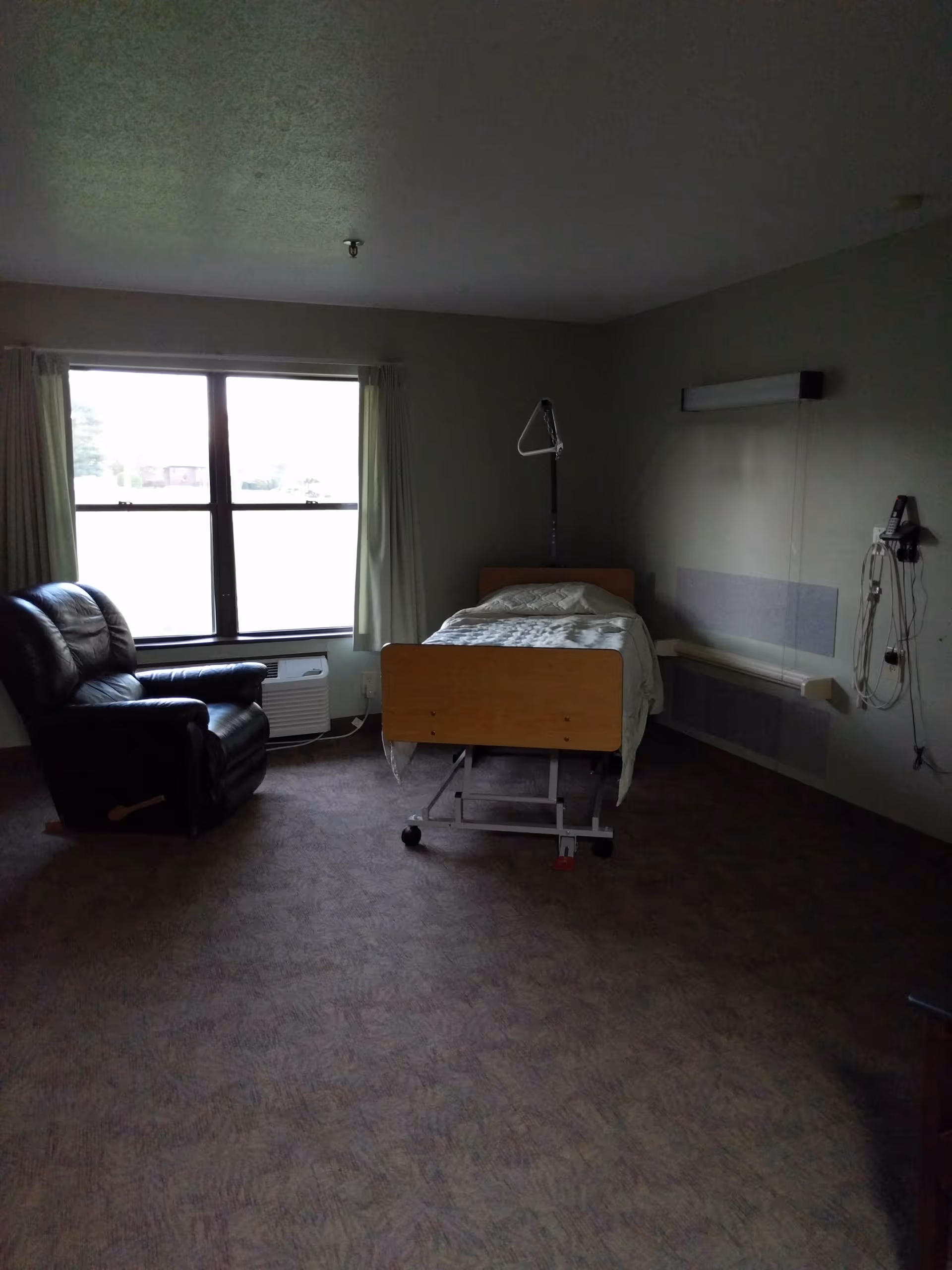 Dimly lit patient room with a bed on rails, a black recliner, and a window.