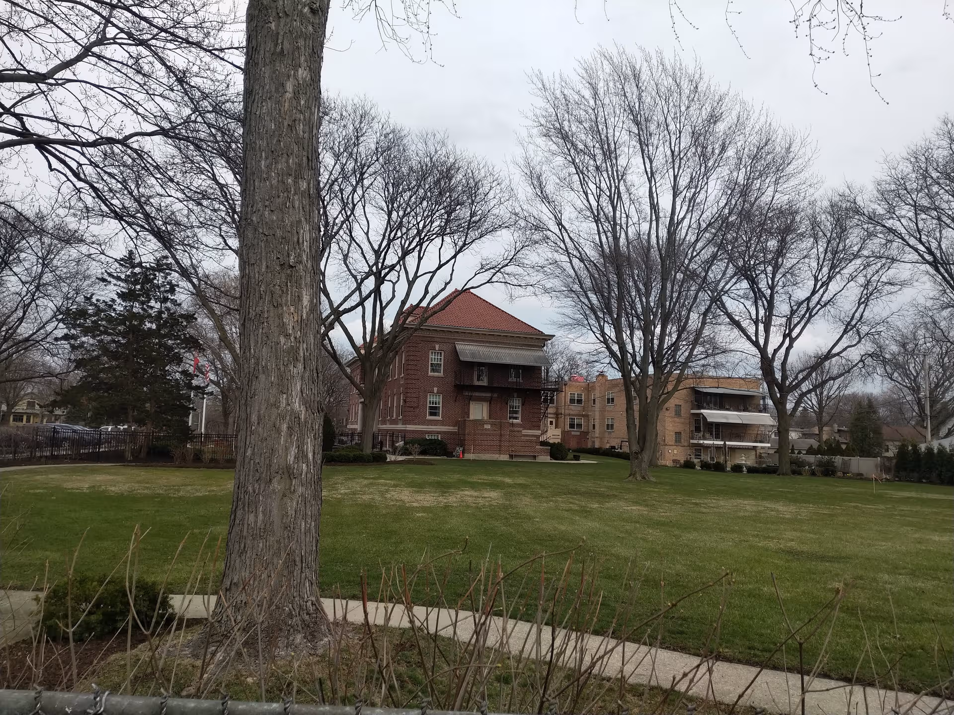 Brick senior living building with balconies on a green lawn under leafless trees.