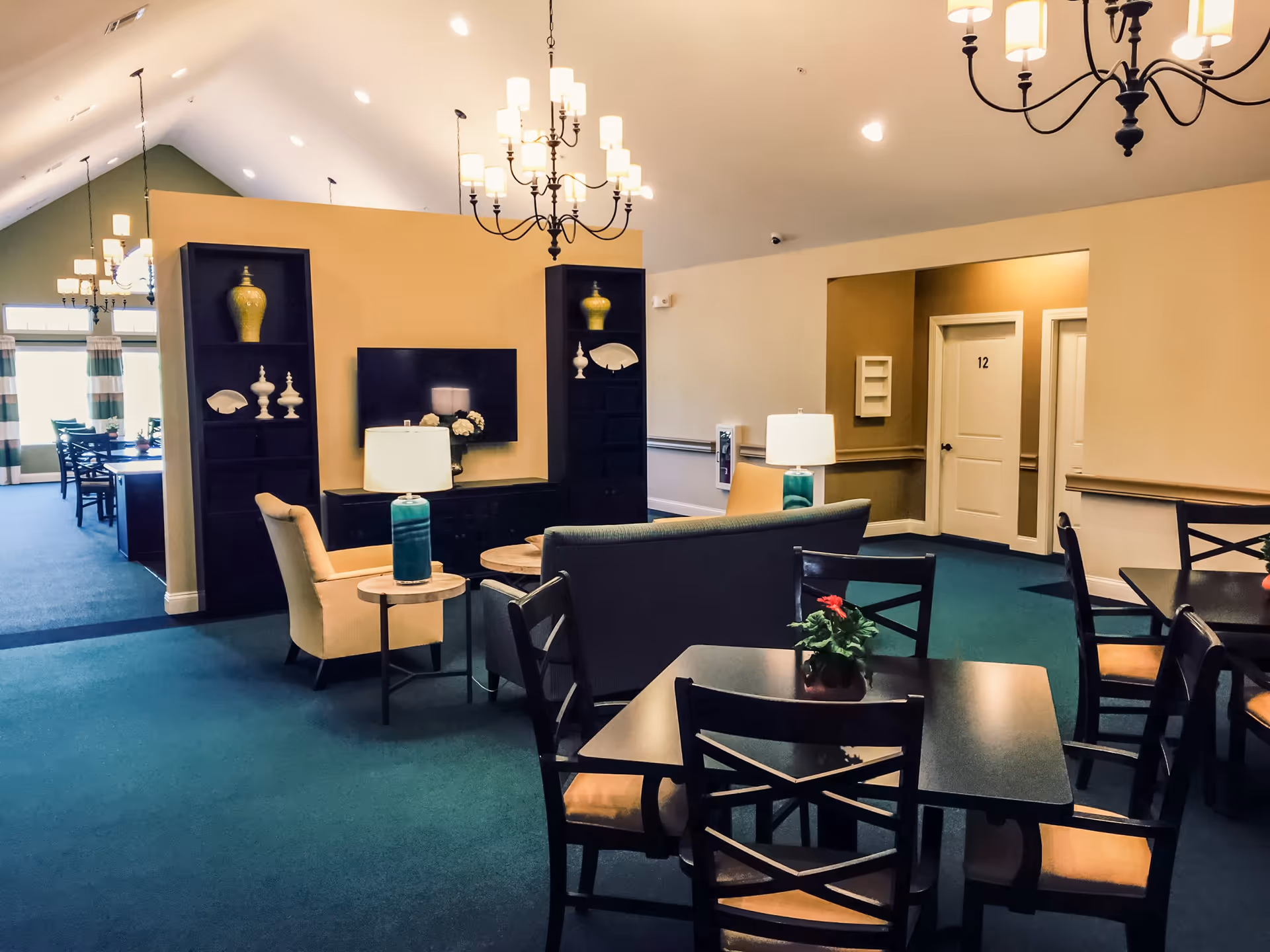 Interior view of a senior living facility common area with blue carpet, beige walls, and high vaulted ceiling. The room features a seating area with a sofa, armchairs, side tables with lamps, and a wall-mounted TV flanked by shelves with decorative items. In the foreground, there are dining tables with chairs and small potted plants. Two white doors are visible in the background, one labeled with the number 12. Multiple chandeliers hang from the ceiling providing warm lighting.