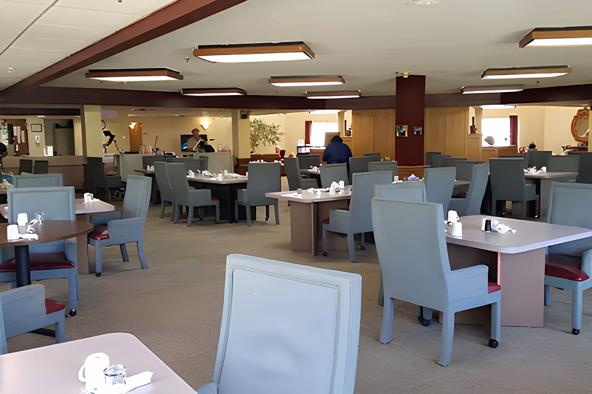 Large dining room with multiple tables and upholstered chairs set for meal service and staff visible in the background.