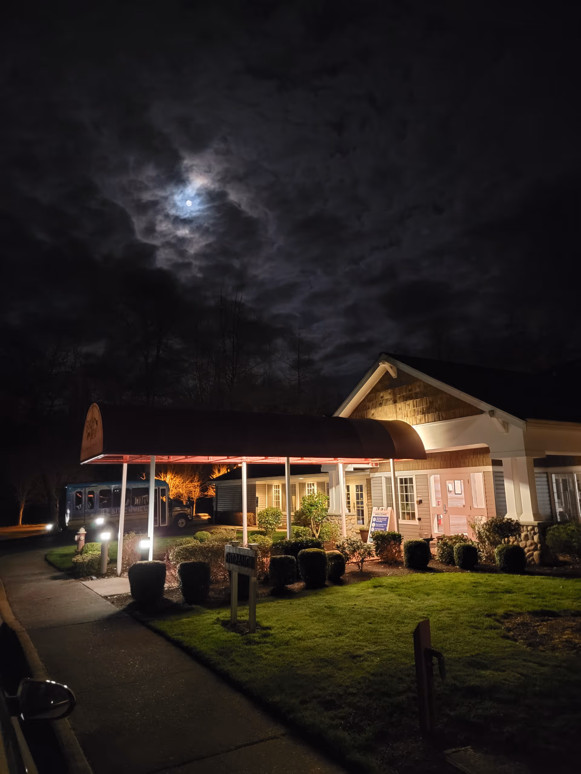 Night exterior of Birchview Memory Care entrance with curved red canopy and a bus nearby.