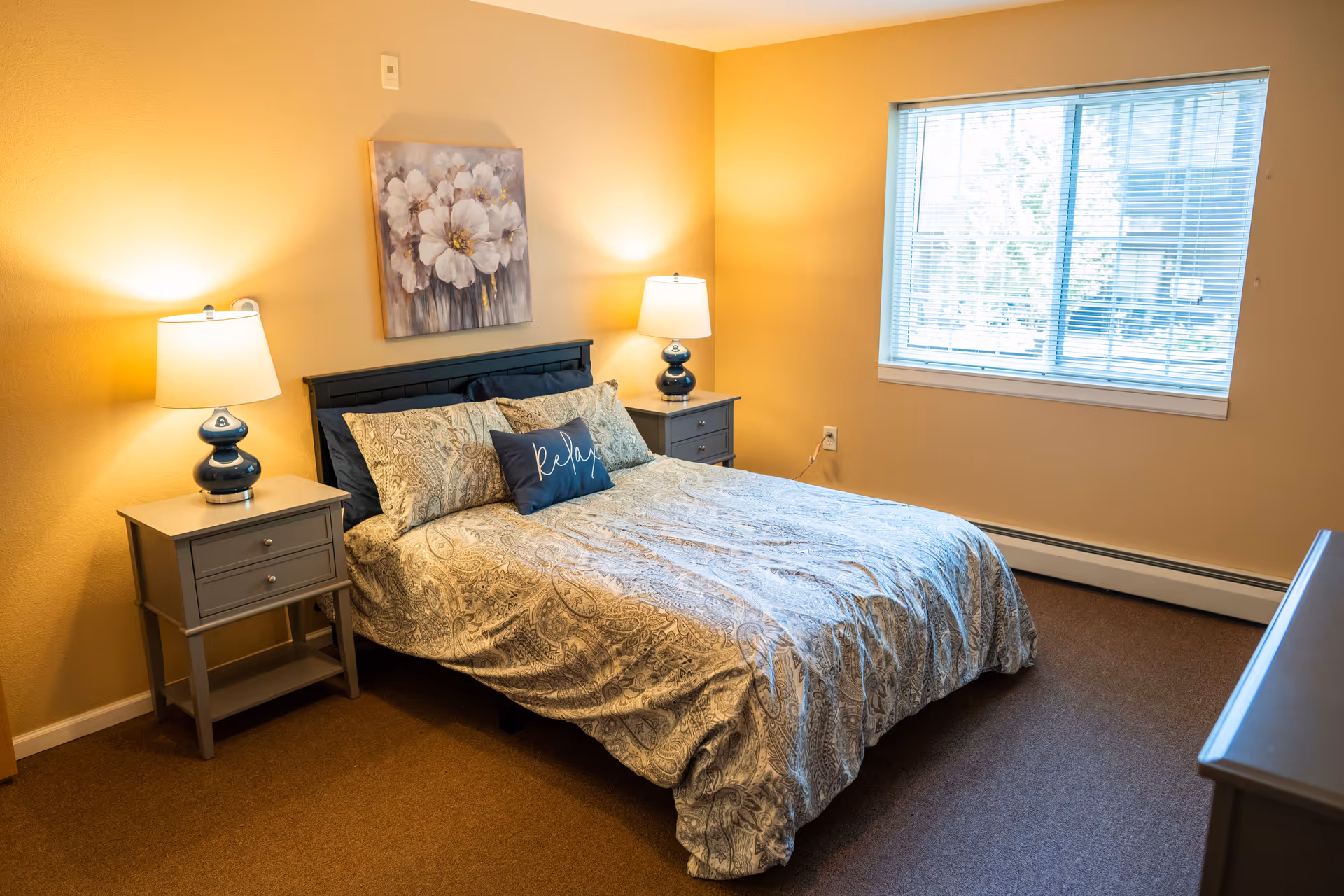 A cozy bedroom with a double bed featuring patterned bedding and a decorative pillow with the word 'Relax'. There are two matching bedside tables with lamps on each side of the bed. A floral painting hangs above the headboard. A window with blinds allows natural light into the room, which has warm yellow walls and brown carpeted flooring.