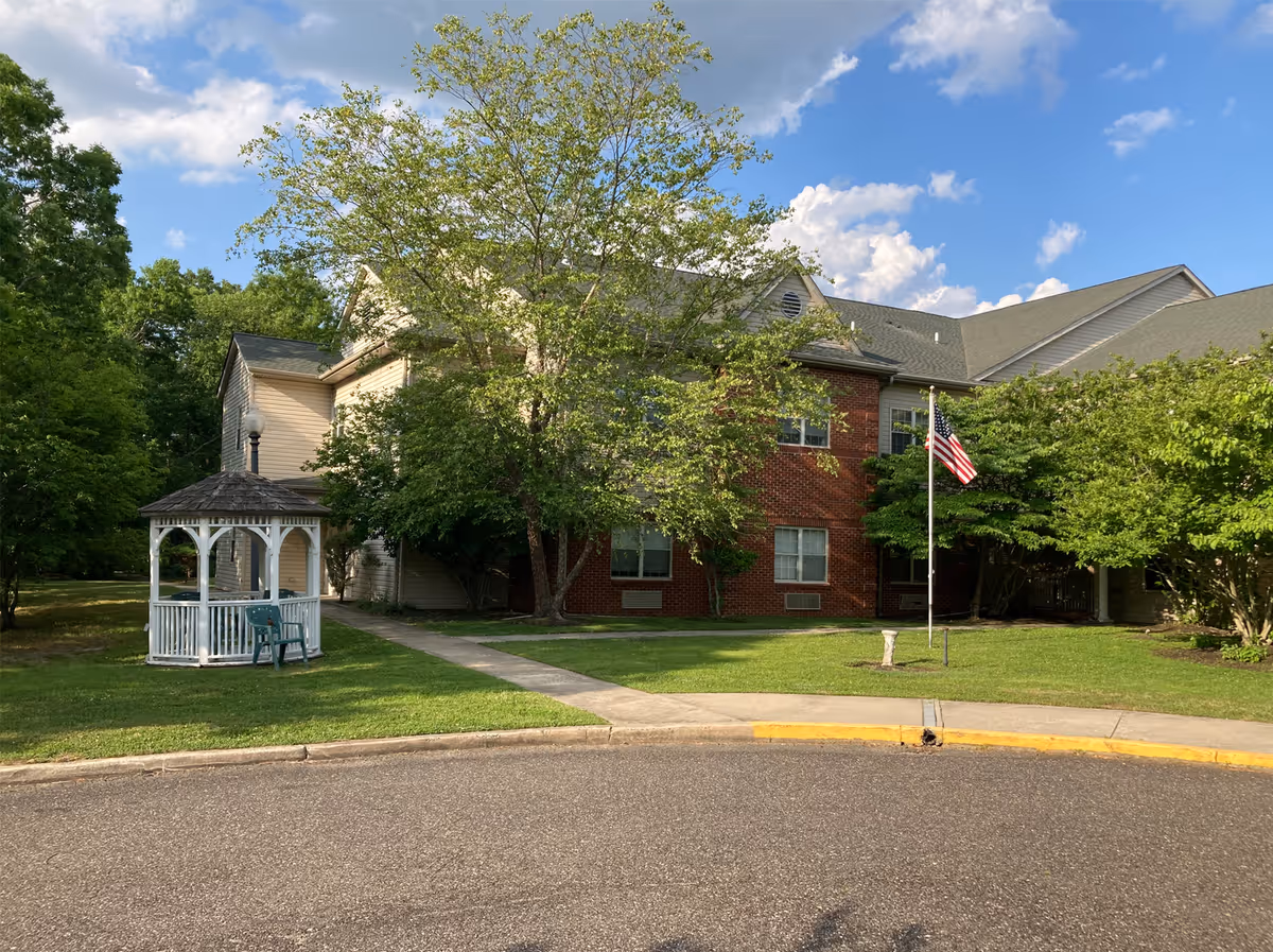 Front view of a brick-and-siding senior living building with a white gazebo, flagpole flying an American flag, and surrounding trees under a blue sky.