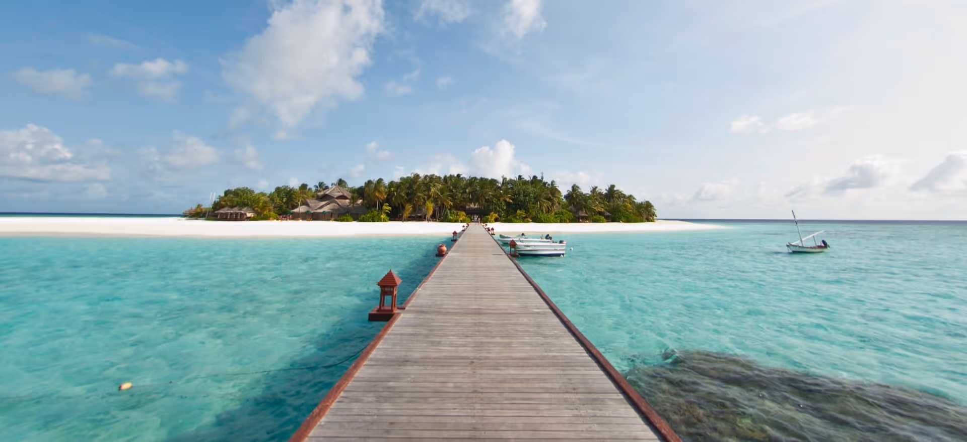 Long wooden pier leads to a tropical island with white sand and turquoise water.