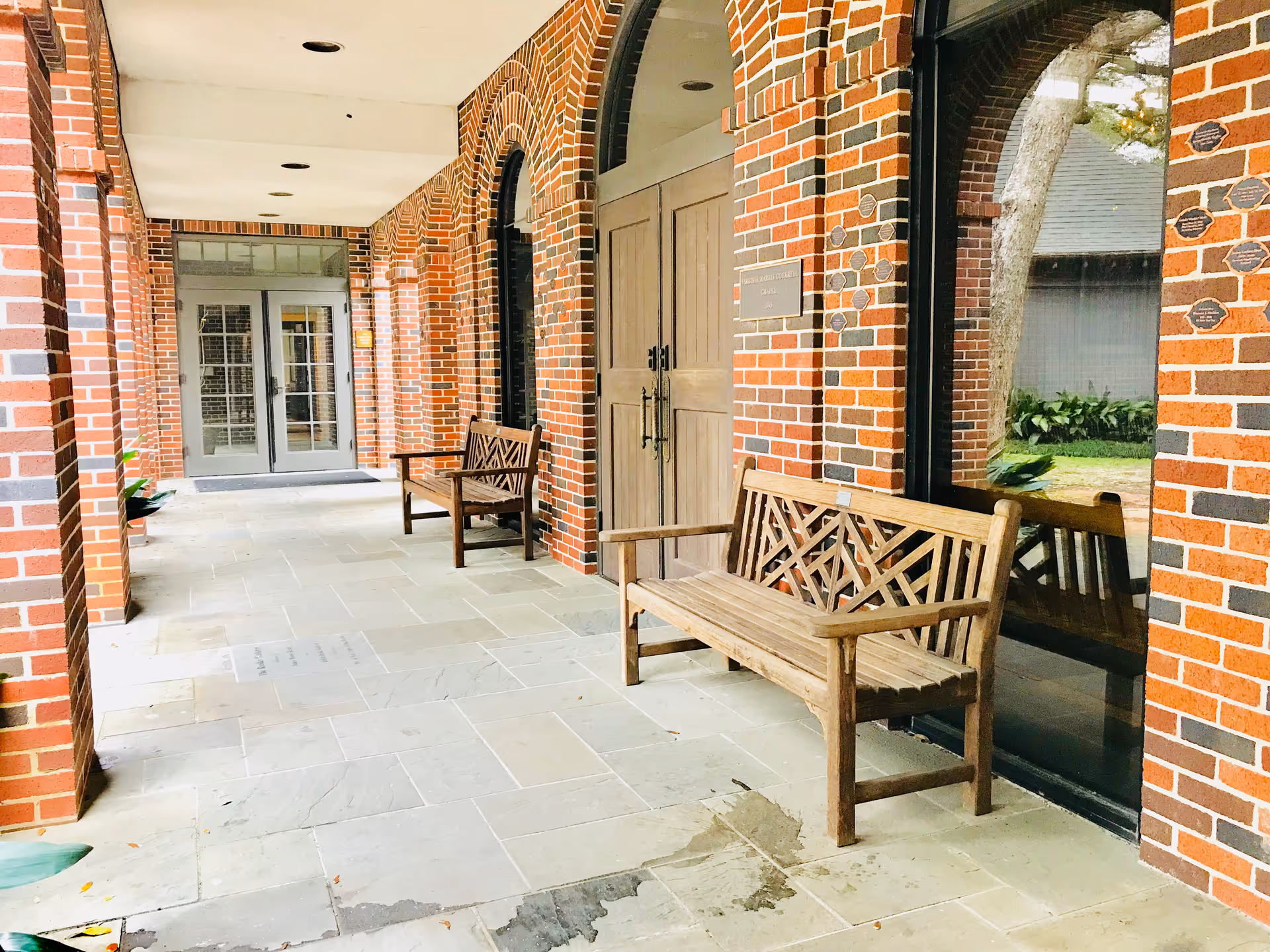 Exterior brick walkway of Houston Hospice with wooden benches and glass doors.