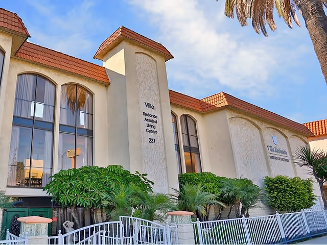 Front exterior of the Villa Redondo assisted living center showing arched windows, a red-tiled roof, palm trees, and a white fence.
