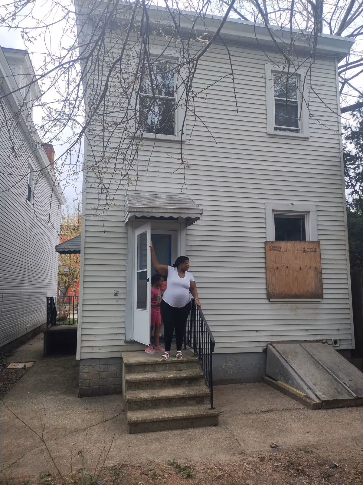 Two people stand on concrete steps outside a two-story beige house with a boarded window.