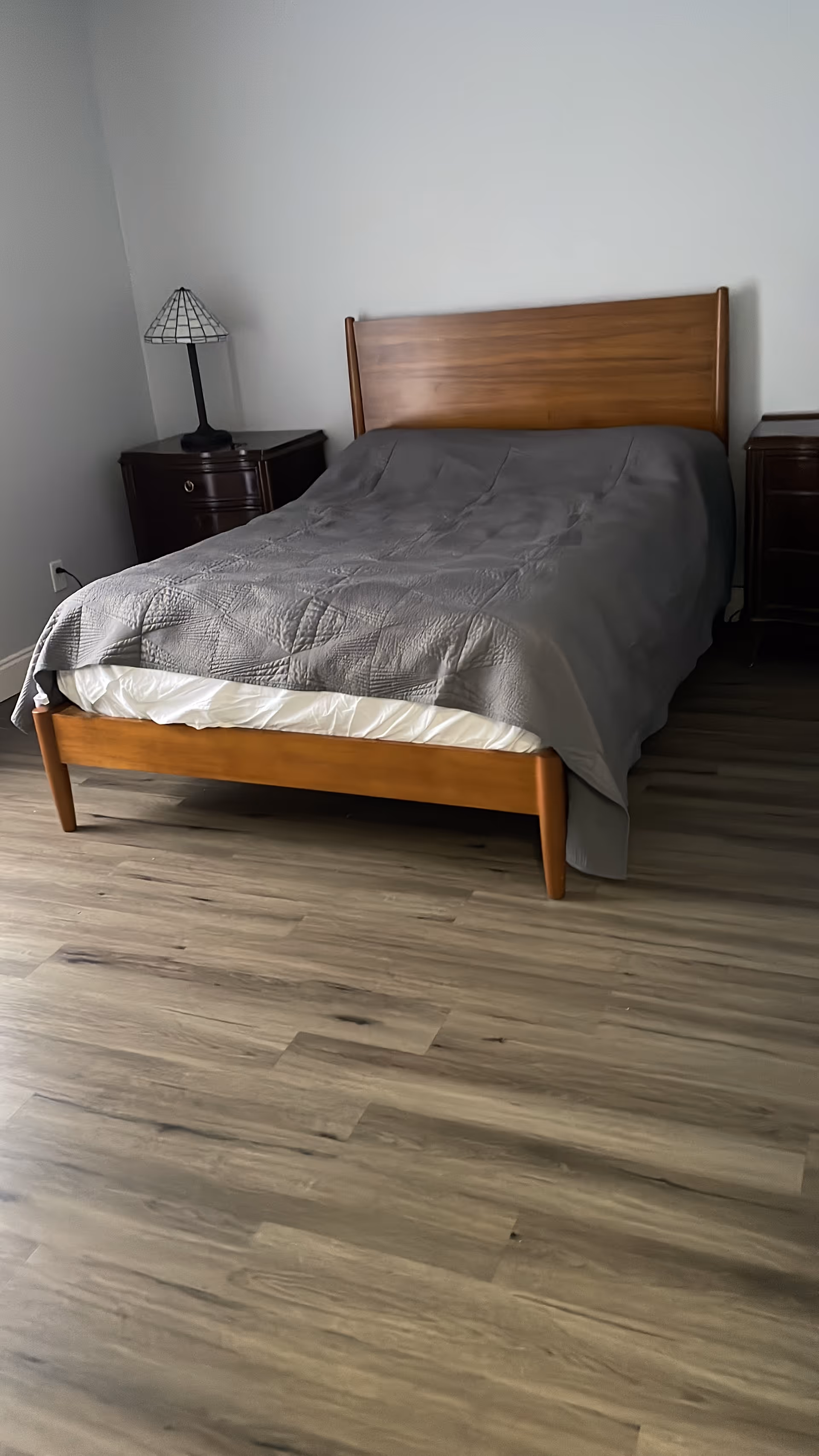 Bedroom with a wooden bed, gray quilt, lamp, and two dark nightstands.