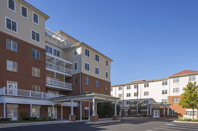 Exterior view of a multi-story senior living facility building with a covered entrance, multiple windows, and a clear blue sky.