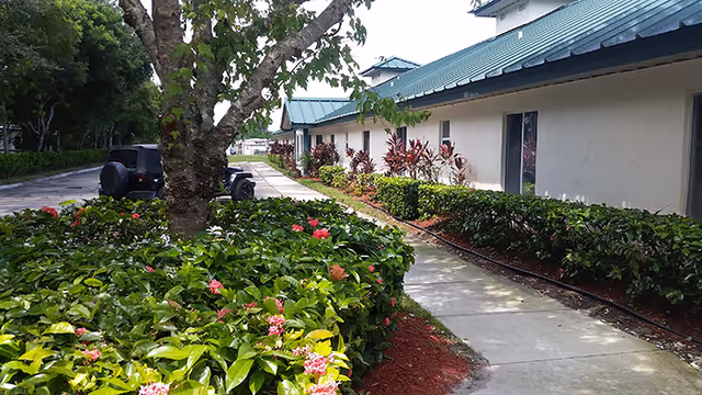 A walkway runs beside a beige assisted living building with manicured hedges and trees.