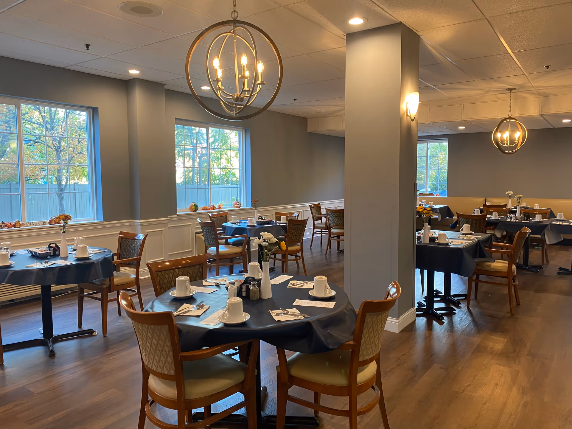 A dining room in a senior living facility with several round tables covered with dark tablecloths. Each table is set with white cups, plates, silverware, and napkins. There are wooden chairs with cushioned seats around the tables. The room has large windows letting in natural light, wood flooring, and modern light fixtures hanging from the ceiling. Some tables have small flower vases as centerpieces.