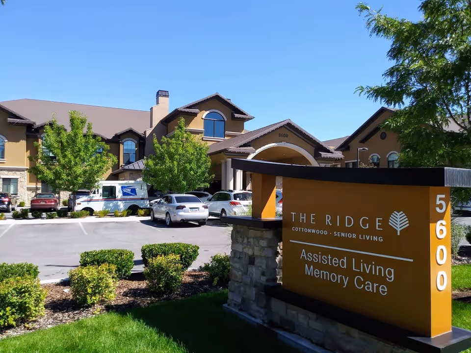 Front exterior and entrance of The Ridge Cottonwood assisted living facility with a large orange sign reading Assisted Living Memory Care and parked cars in the lot.