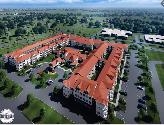 Aerial view of a large senior living facility with multiple connected buildings featuring red roofs and white walls, surrounded by parking lots and green landscaped areas with trees and grass.