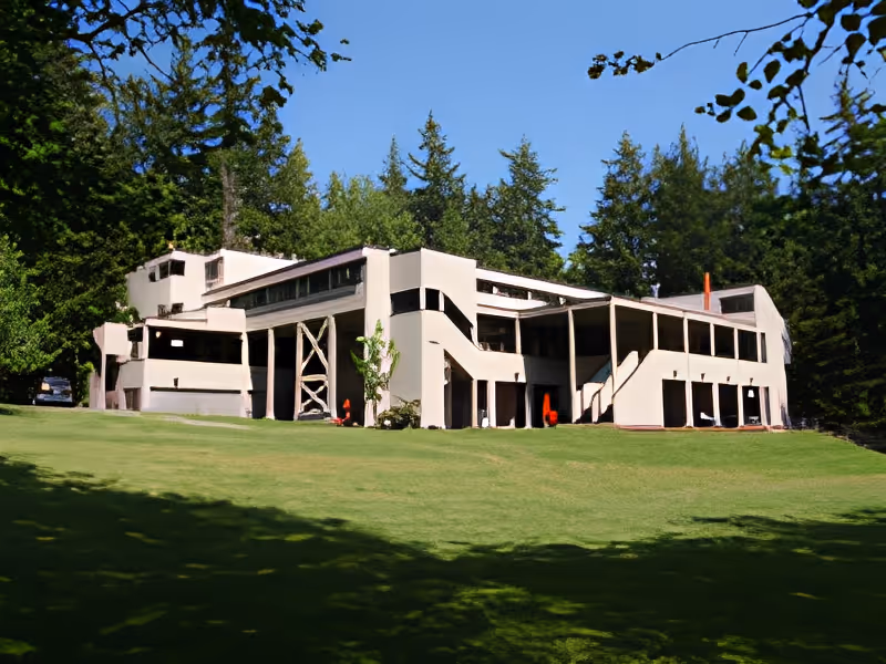 Modern multi-level building with covered walkways and exterior staircases on a large grassy lawn surrounded by trees.