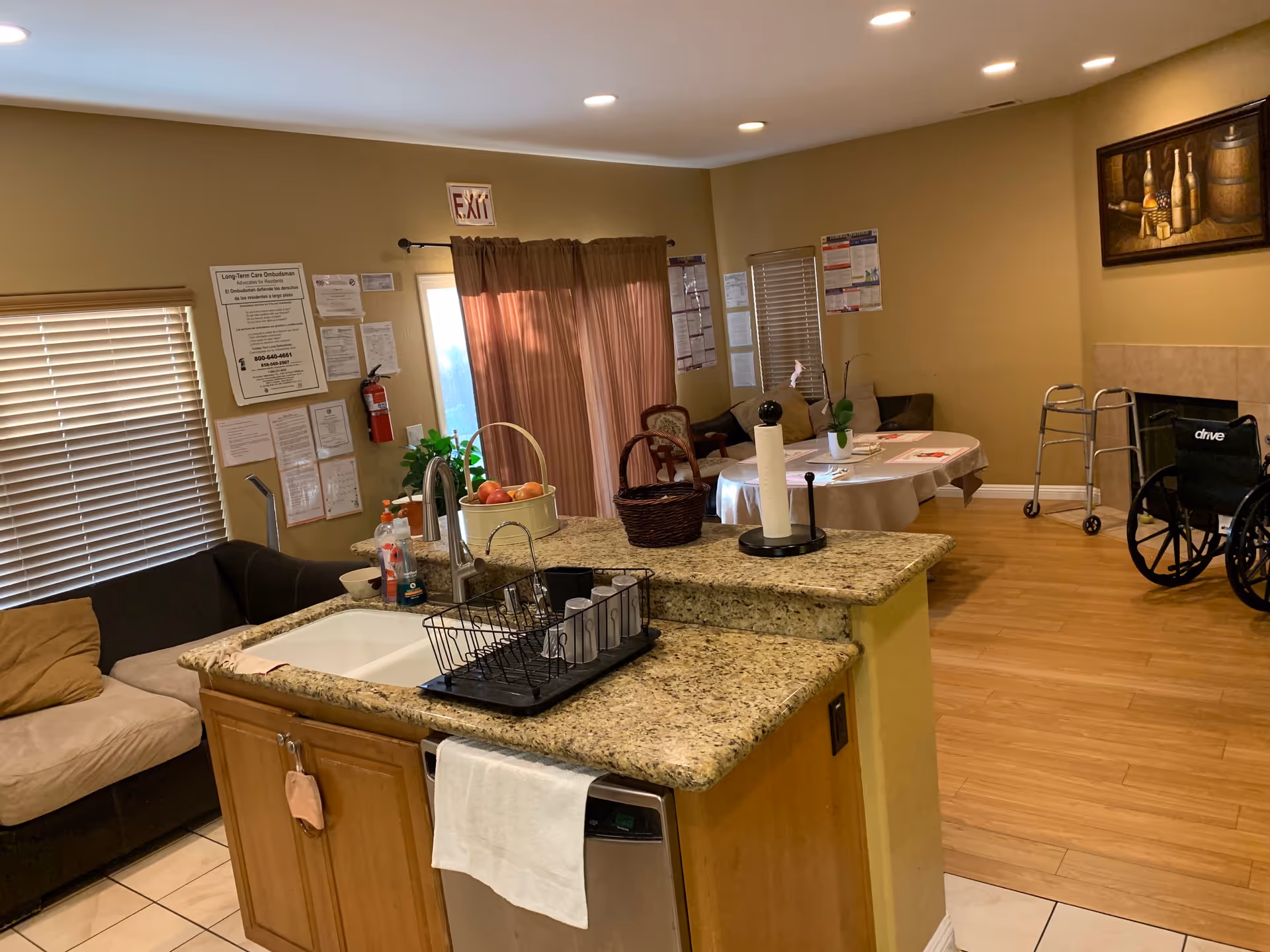 Interior view of a senior living facility showing a kitchen island with a sink, dish rack, and paper towel holder. In the background, there is a dining table with chairs, a couch, a wheelchair, a walker, and various notices on the walls. The room has wooden flooring and beige walls with a painting above a fireplace.