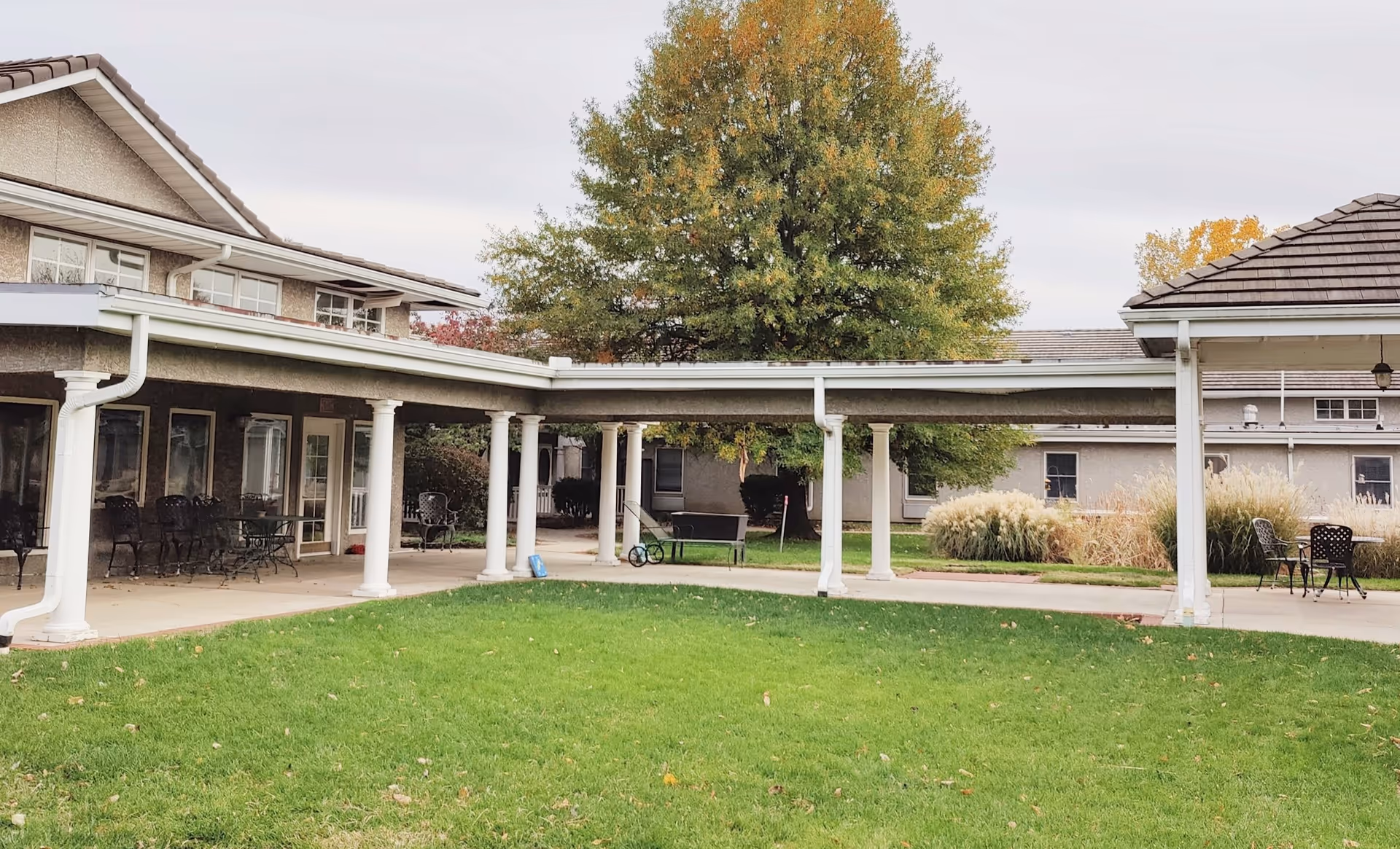 Outdoor courtyard area of Colonial Oaks Senior Living at Leawood featuring a covered walkway supported by white columns, green grass lawn, patio seating with tables and chairs, and trees with autumn foliage.