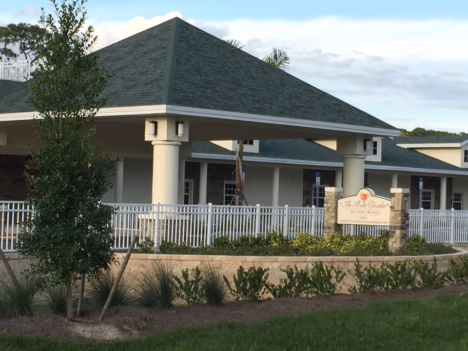Front entrance of The Rose Garden of Fort Myers building with a covered portico, white fence, landscaping, and facility sign.