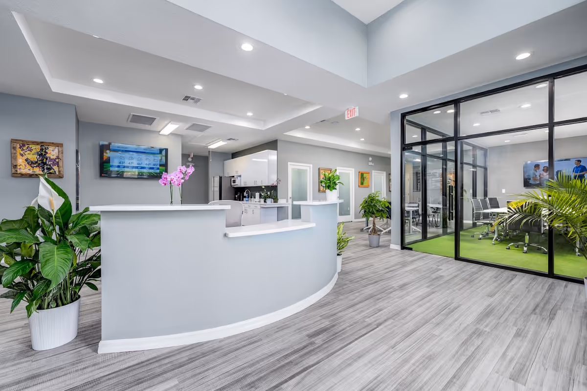 Modern senior living lobby with curved reception desk, plants, and glass-walled meeting room.