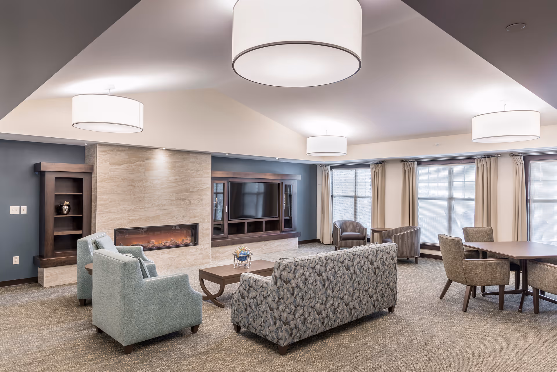 A spacious senior living community common area with a modern fireplace set in a stone wall, a flat-screen TV mounted above a wooden cabinet, and comfortable seating including patterned sofas and armchairs. Large windows with beige curtains allow natural light to fill the room, and there is a dining table with chairs on the right side. The ceiling features three large round light fixtures.