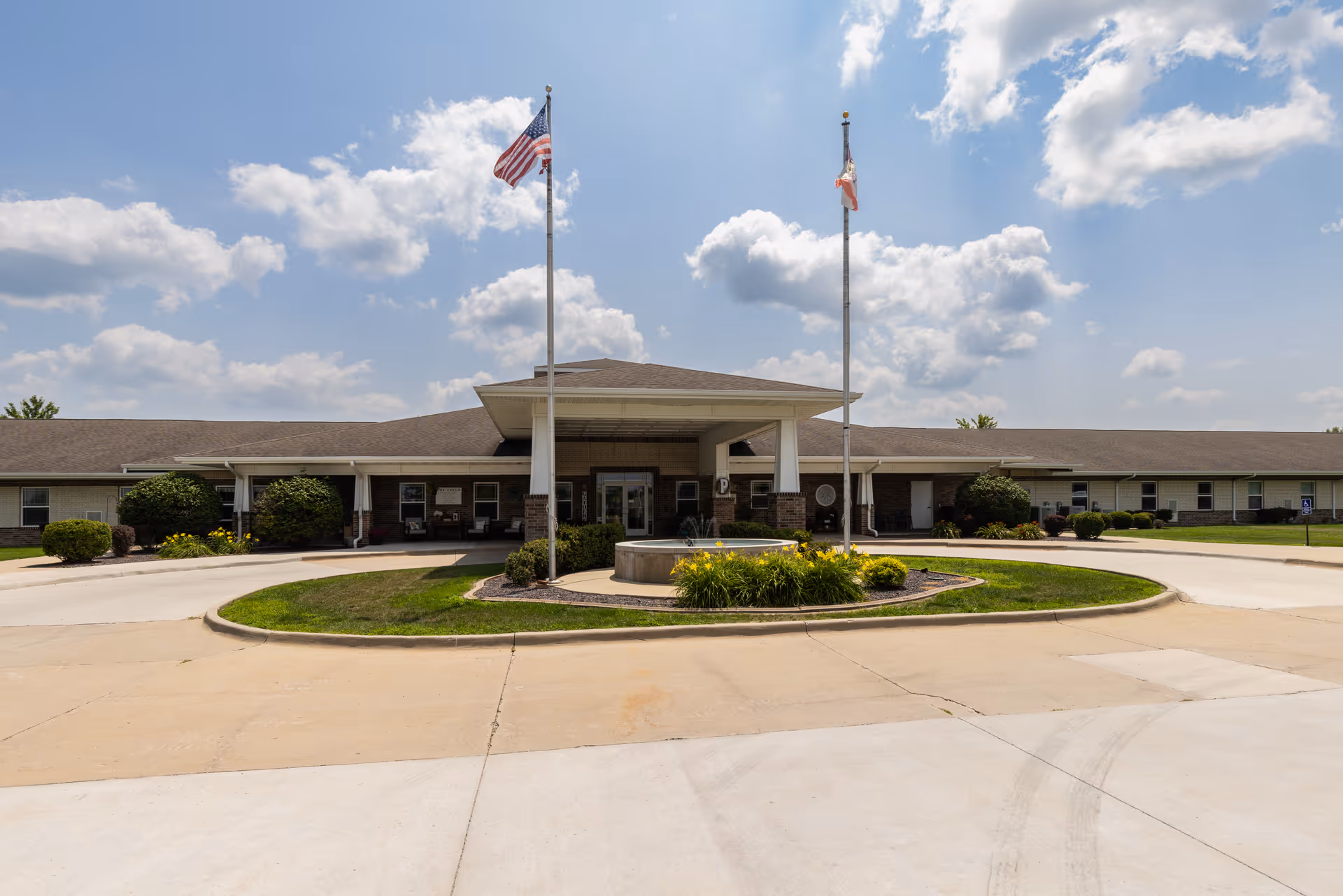 Front exterior view of Addington Place of Mount Pleasant building with two flagpoles displaying the American flag and another flag, a circular driveway with a central landscaped area featuring a small fountain and yellow flowers, under a partly cloudy blue sky.