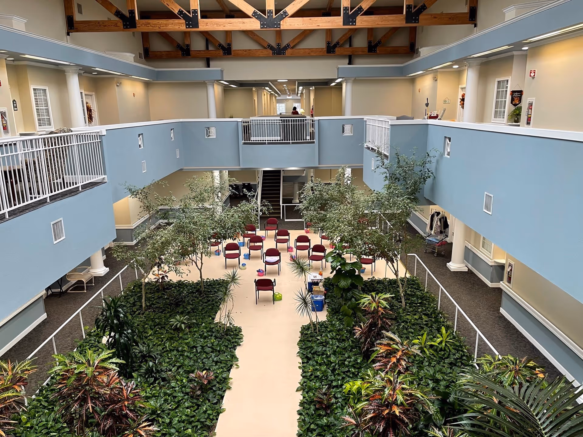 Interior view of a spacious assisted living facility atrium with a central walkway lined with green plants and small trees. Several red chairs are arranged in rows at the far end of the walkway. The upper level features blue walls with white railings and exposed wooden beams on the ceiling.