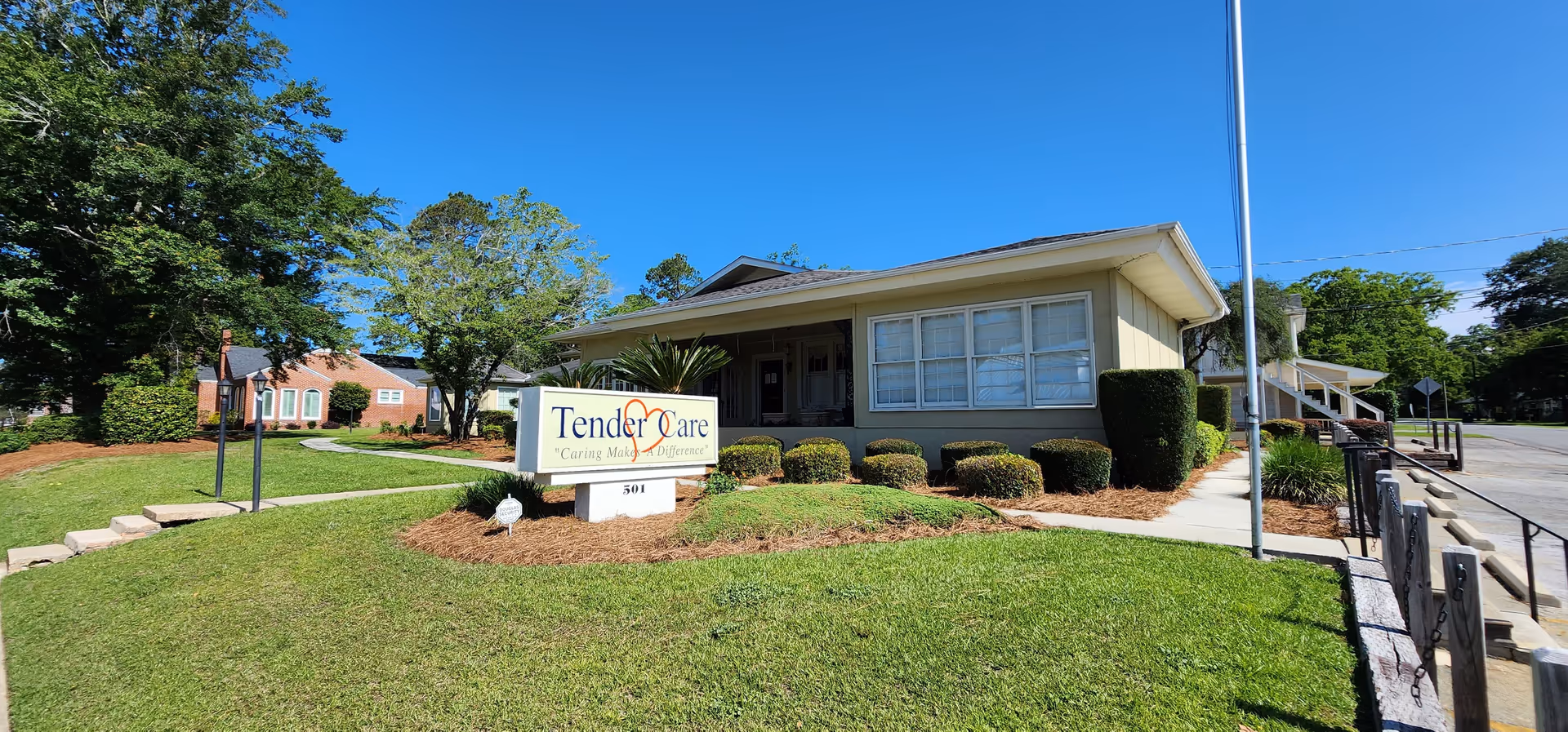 Single-story Tender Care facility exterior with sign, trimmed shrubs, and bright blue sky.