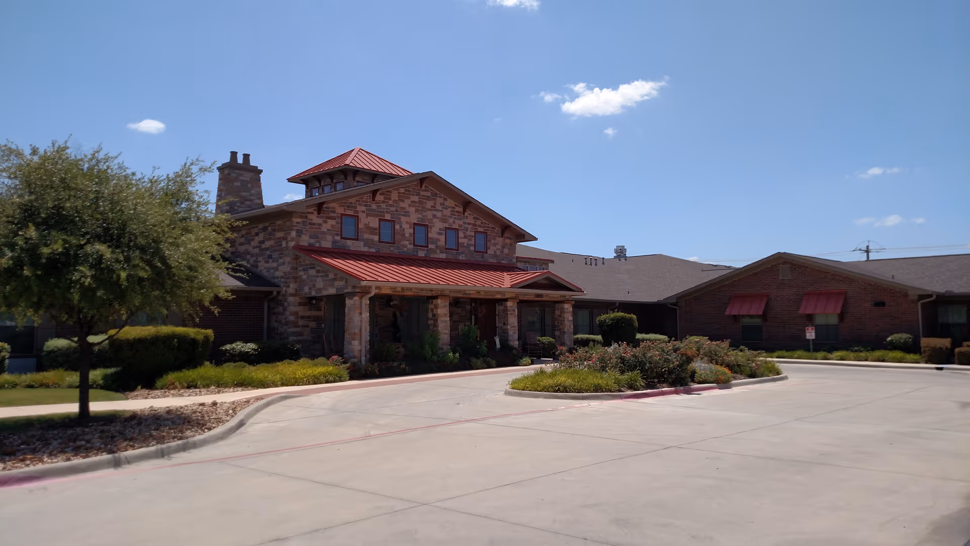 Stone-brick senior living facility front with driveway, landscaped island, and a clear blue sky.