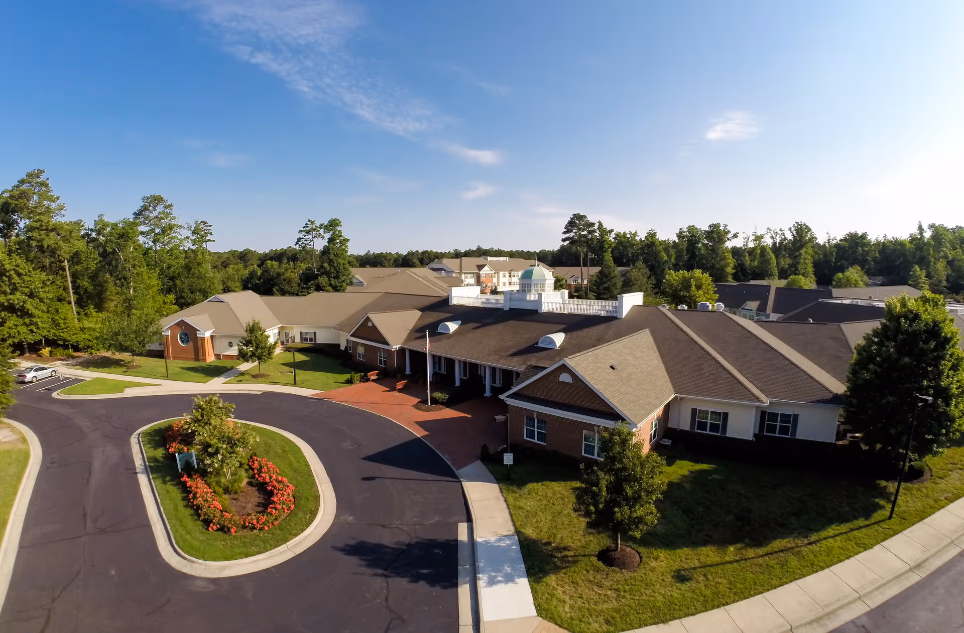 Aerial view of a senior living facility named Lake Prince Woods, showing a large building with multiple roof sections, surrounded by trees and greenery. There is a circular driveway with landscaped flower beds and a few parked cars. The sky is clear and blue.