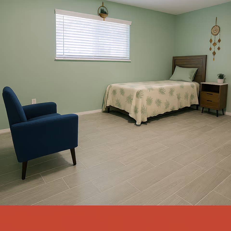 A pastel-green bedroom with a single bed, palm-pattern bedspread, blue chair, and nightstand.