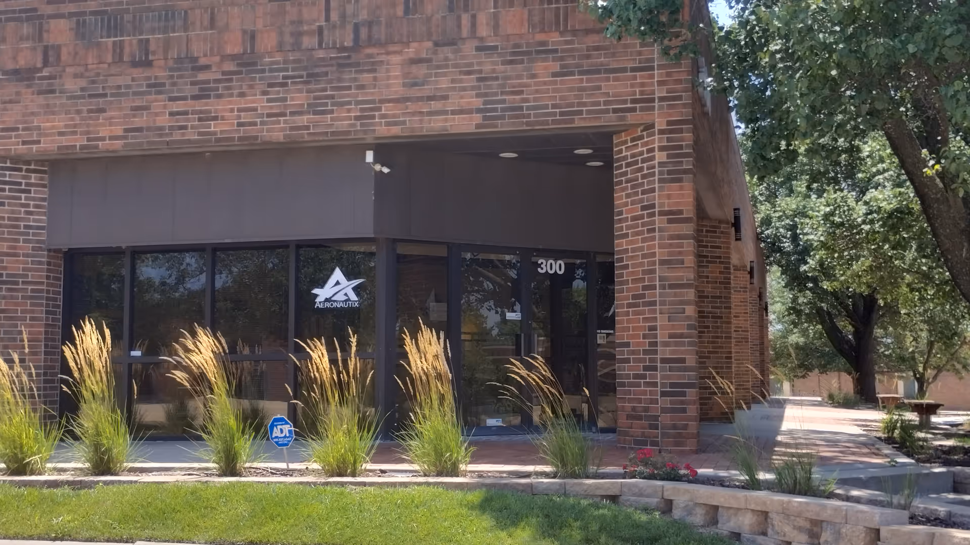 Brick building entrance with glass doors, landscaped walkway and trees nearby.