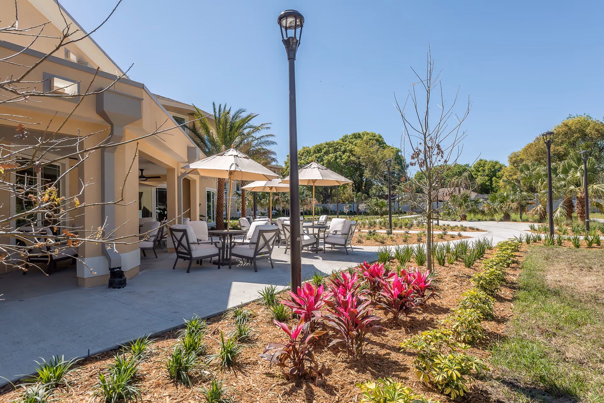 Outdoor patio and landscaped courtyard with tables, umbrellas, lounge chairs and a building facade under a clear sky.