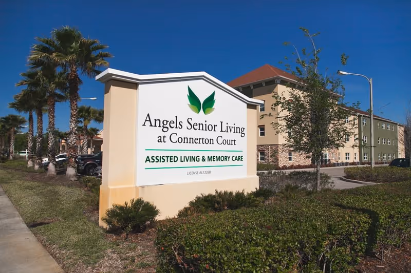 Entrance sign for Angels Senior Living at Connerton Court in front of a multi-story assisted living building and palm trees.