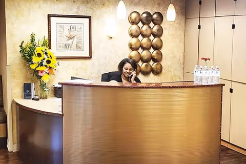 Reception area with a curved wooden desk where a woman is sitting and talking on the phone. Behind the desk, there is a decorative wall piece with round metallic elements, a framed clock on the wall, and a vase with sunflowers and other flowers. Several bottles of water are placed on the desk.