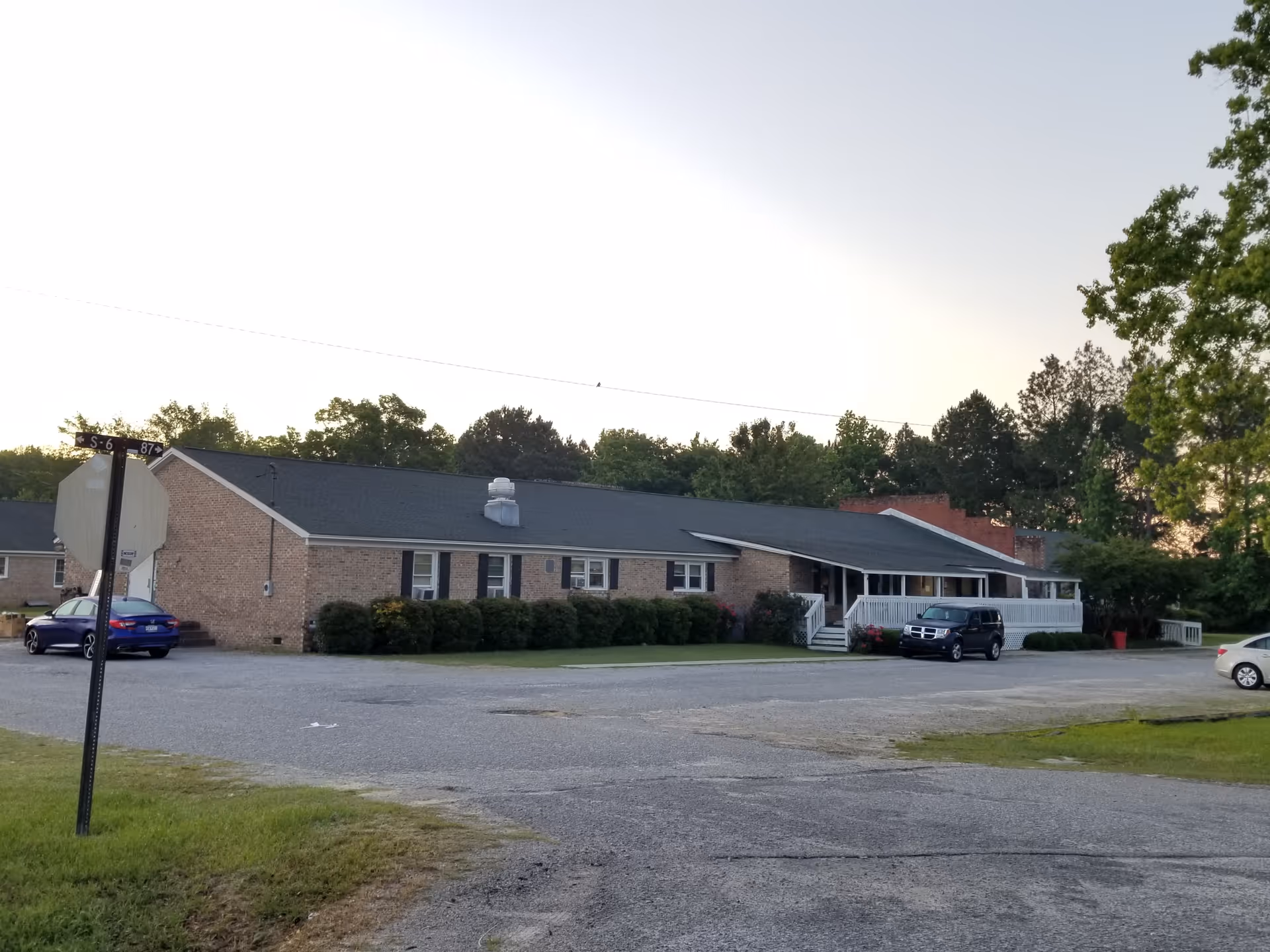 Single-story brick facility with parking and shrub-lined front, viewed from a gravel lot.