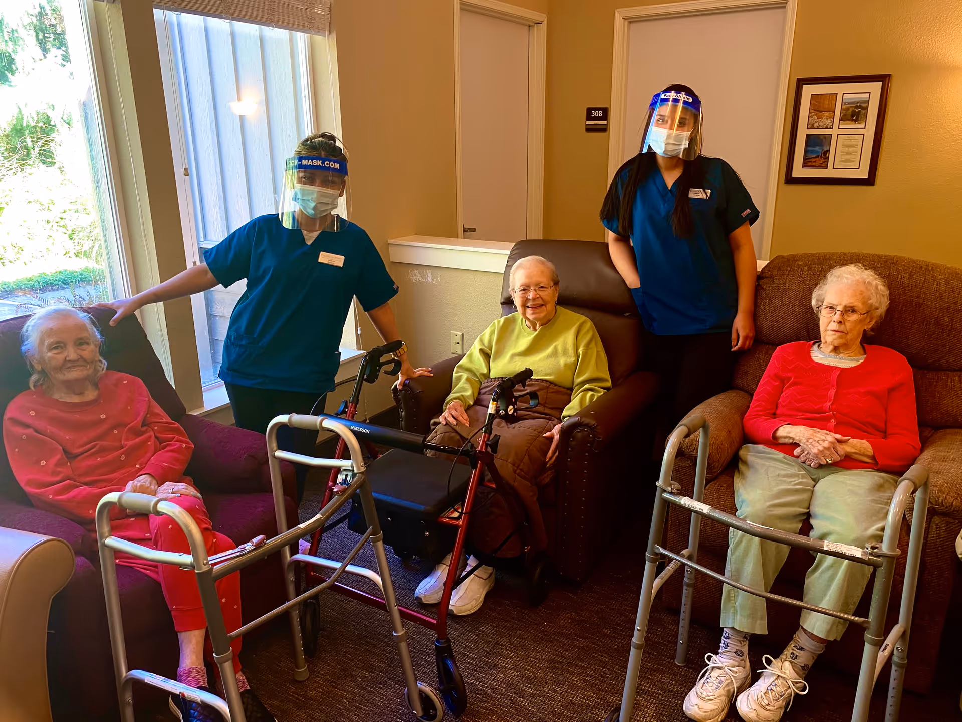 Three elderly residents seated with walkers in a common room alongside two masked caregivers wearing face shields.