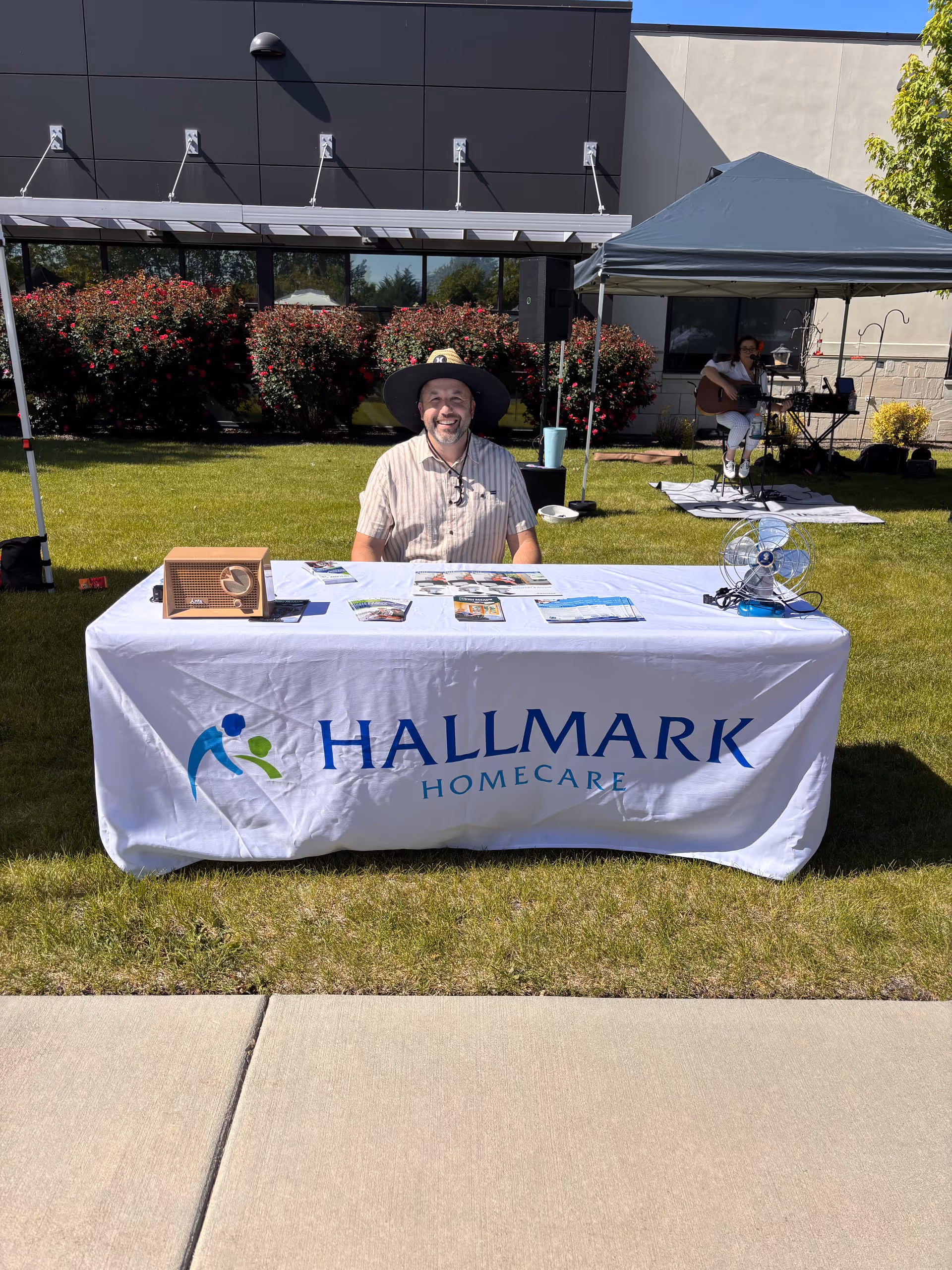 Smiling man sits at a Hallmark Homecare table outdoors; tent with person in background.