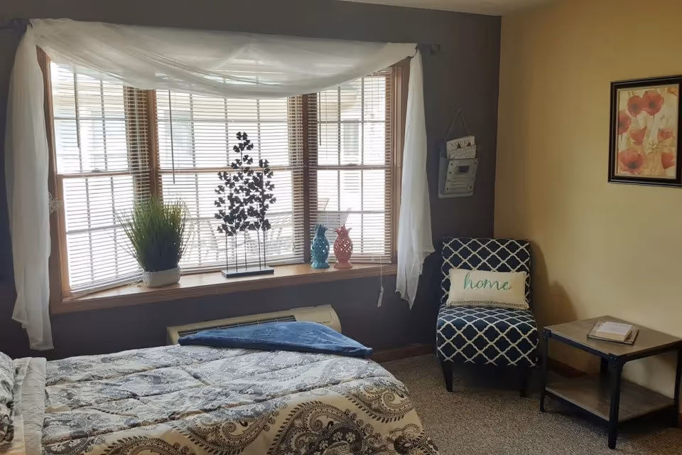 A cozy bedroom with a bed covered in a patterned comforter, a large bay window with blinds and sheer white curtains, a decorative plant and two ceramic pineapples on the window sill, a patterned armchair with a pillow that says 'home', a small side table with a book, and a framed floral picture on the wall.