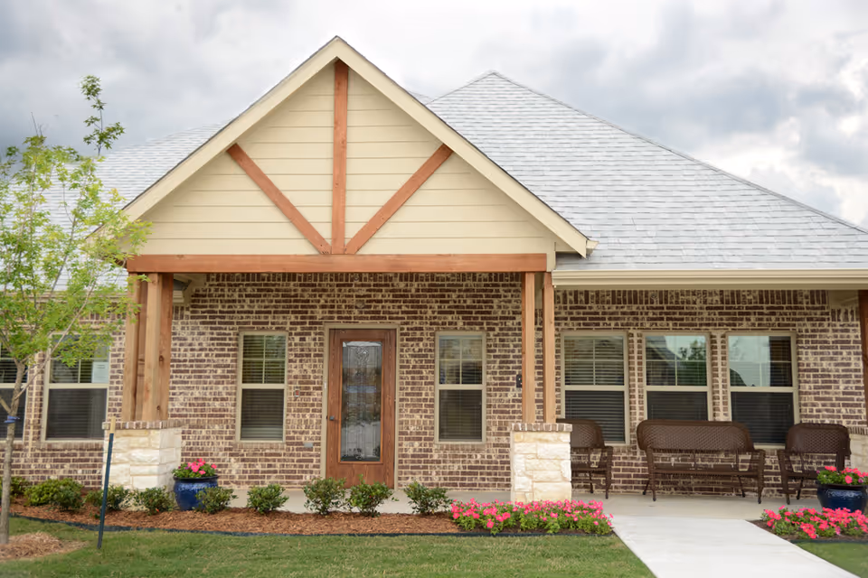 Front exterior view of a single-story brick building with a covered porch supported by wooden beams. The porch has a wooden door with glass panels and several windows with blinds. There are benches on the porch and flower pots with pink flowers on either side of the walkway leading to the door. A small tree and neatly trimmed bushes are in the landscaped area in front of the building.