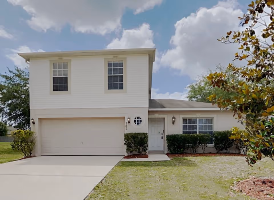 Two-story beige building with attached garage, front entry, and trimmed shrubs.