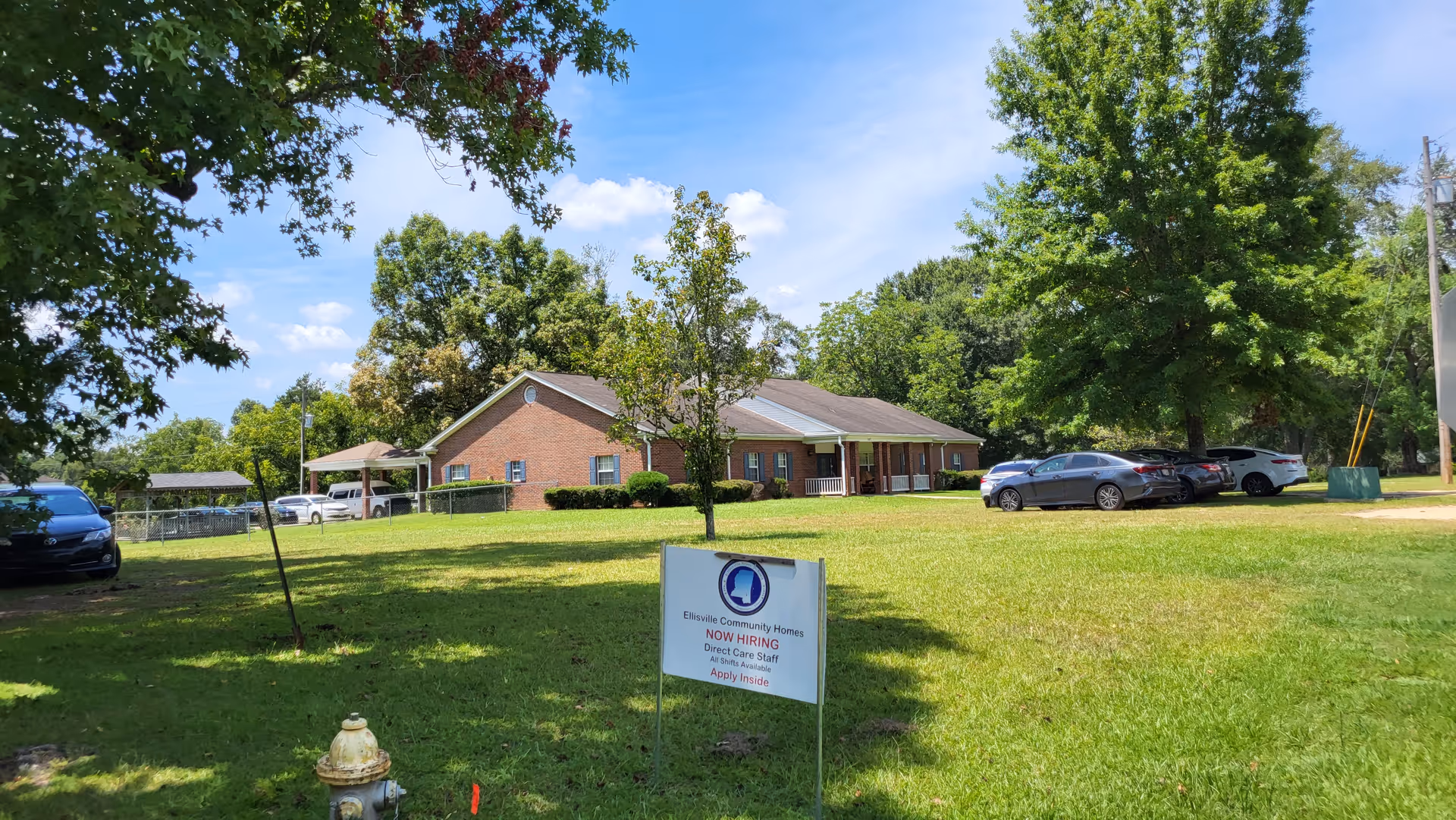 Exterior view of a brick senior living facility with a grassy lawn, trees, and parked cars.