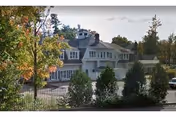 White multi-story nursing center with dormer windows, surrounded by trees and a fence.