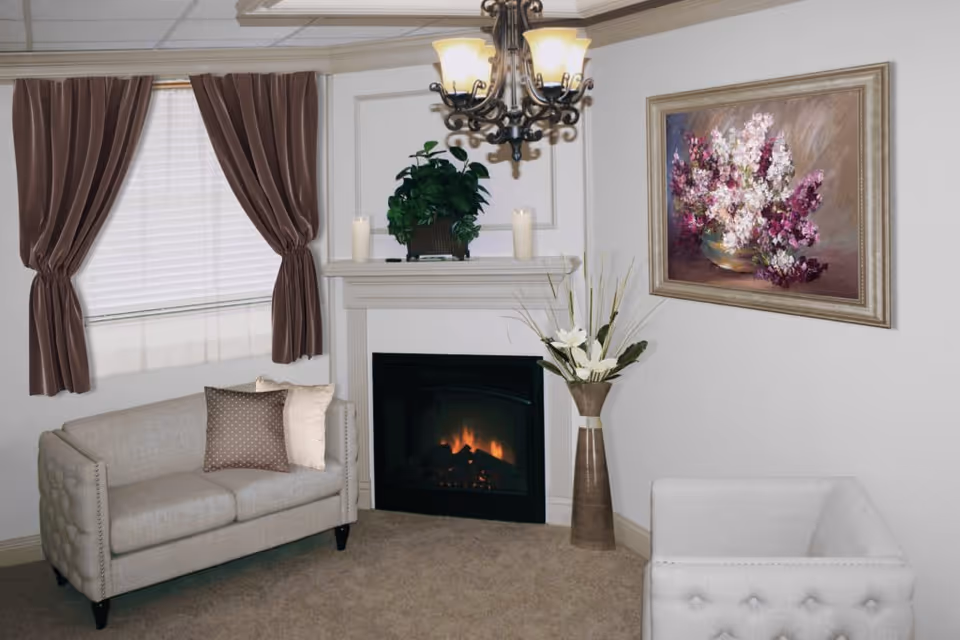A cozy living room with a beige loveseat and a white armchair, a fireplace with a small fire burning, a plant and two candles on the mantel, a tall vase with white flowers, brown curtains on the window, a chandelier hanging from the ceiling, and a framed painting of purple and white flowers on the wall.
