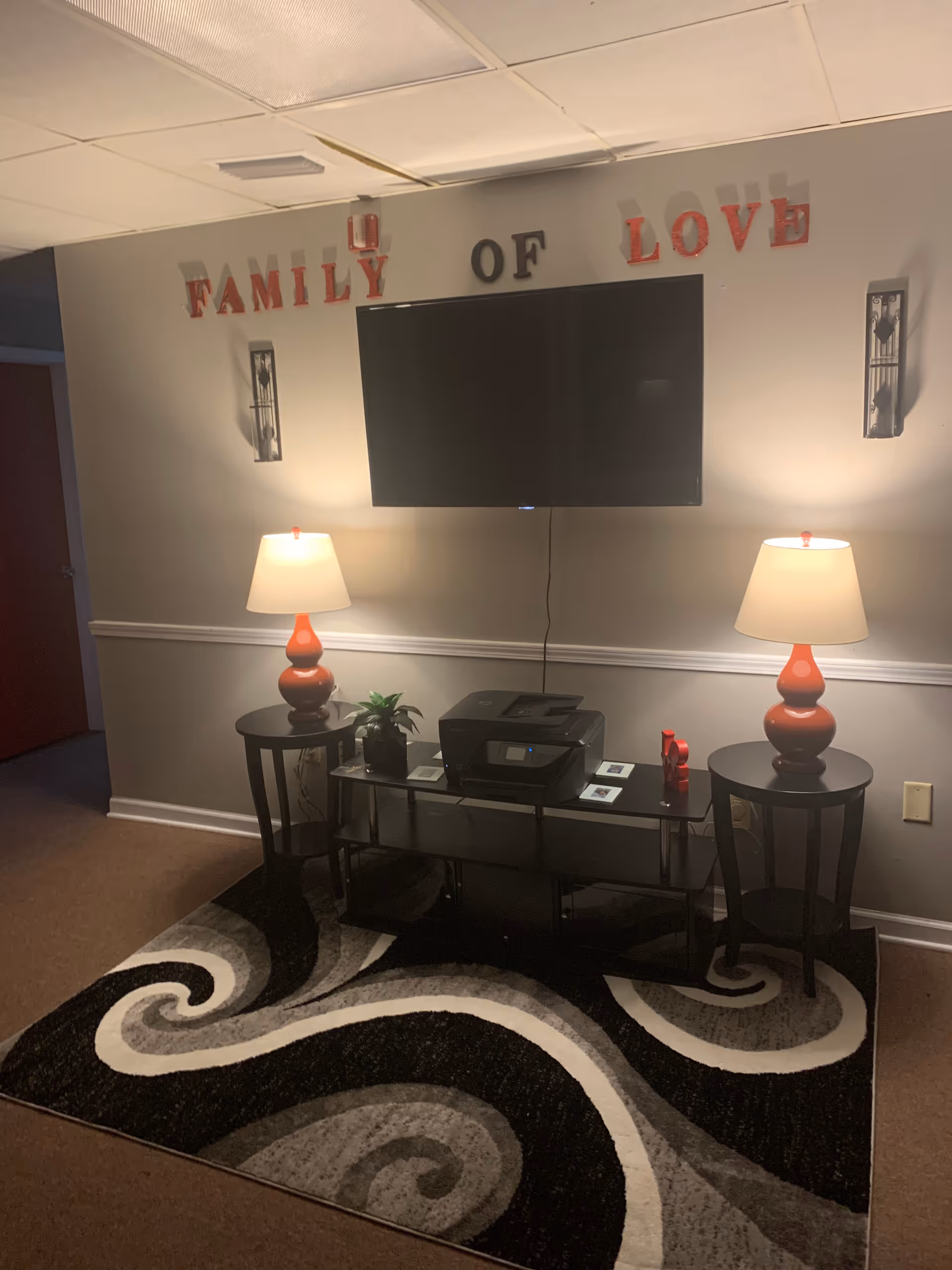 A living room area with wall-mounted TV, two red lamps, console table, and swirling rug.