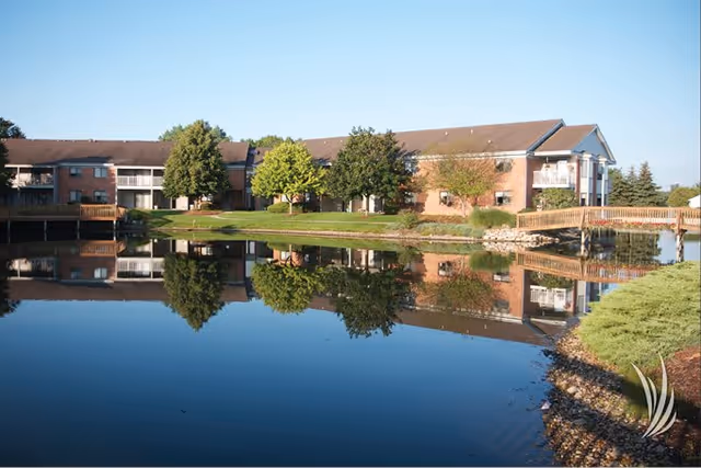 Brick retirement community building and trees reflected in a calm pond under a clear blue sky.