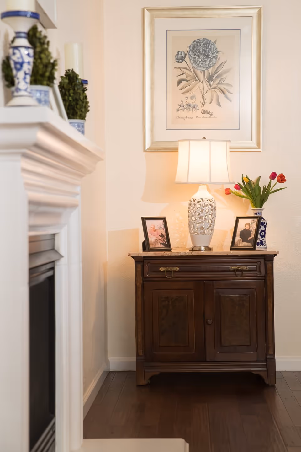 A cozy corner of a living room featuring a wooden cabinet with two framed photos, a white decorative table lamp, and a vase with colorful tulips. Above the cabinet hangs a framed botanical print. To the left, part of a white fireplace mantel is visible with blue and white decorative items and greenery on top.